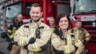 En mannlig og en kvinnelig brannkonstabel står med ryggen mot hverandre og smiler mot kamera
