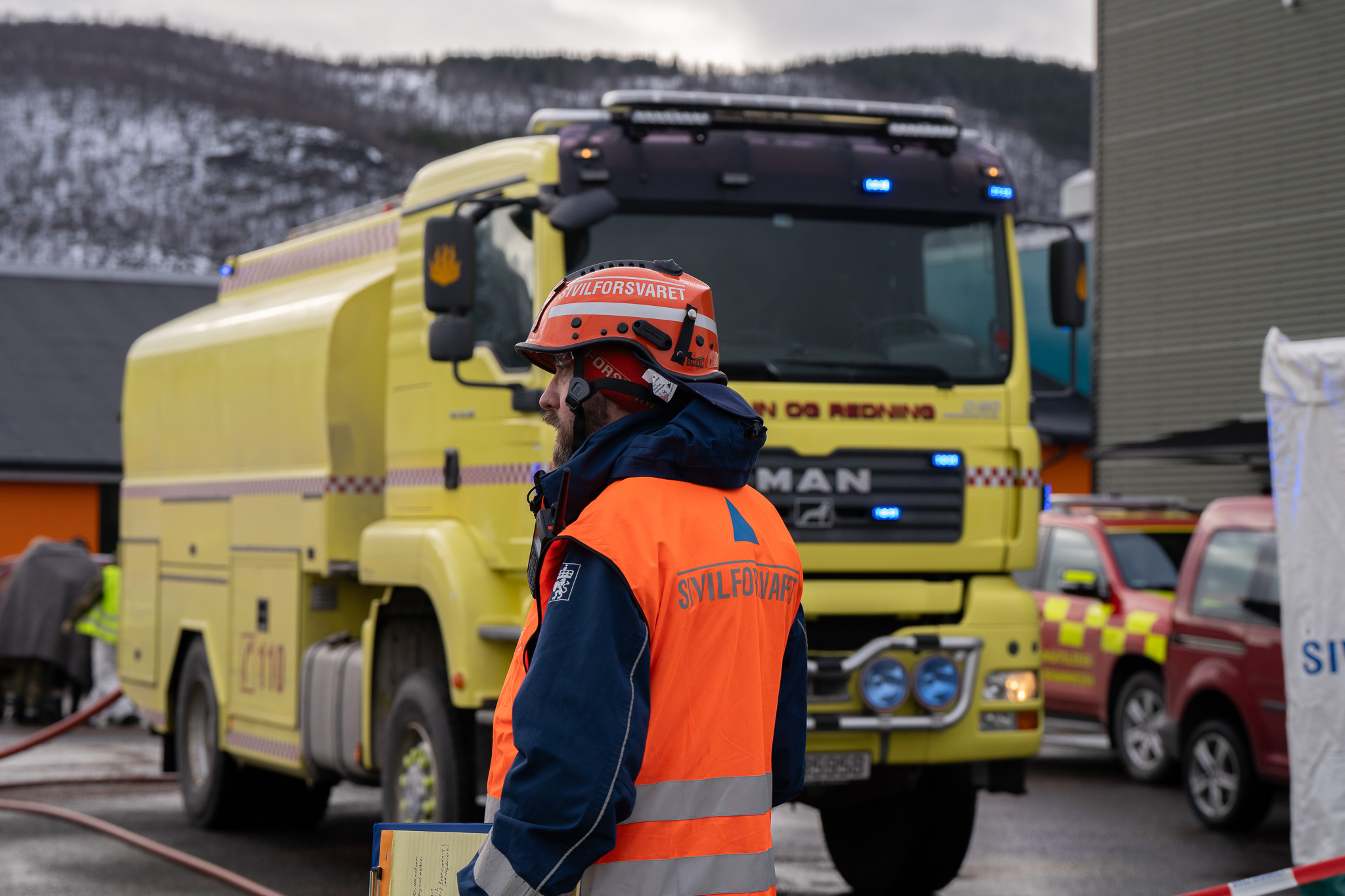 En mann i Sivilforsvarets uniform står i forgrunn. I bakgrunn er det en gul brannbil.