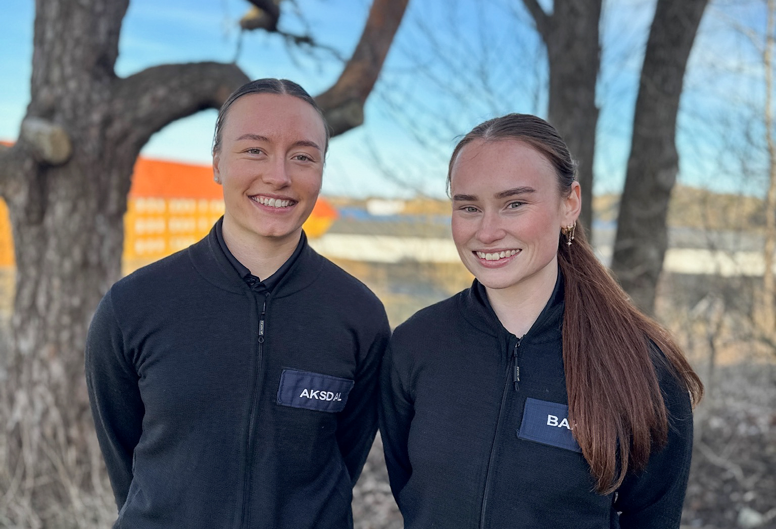 To kvinnelige studenter står ute og smiler til kamera. Bak dem er det trær og blå himmel. 