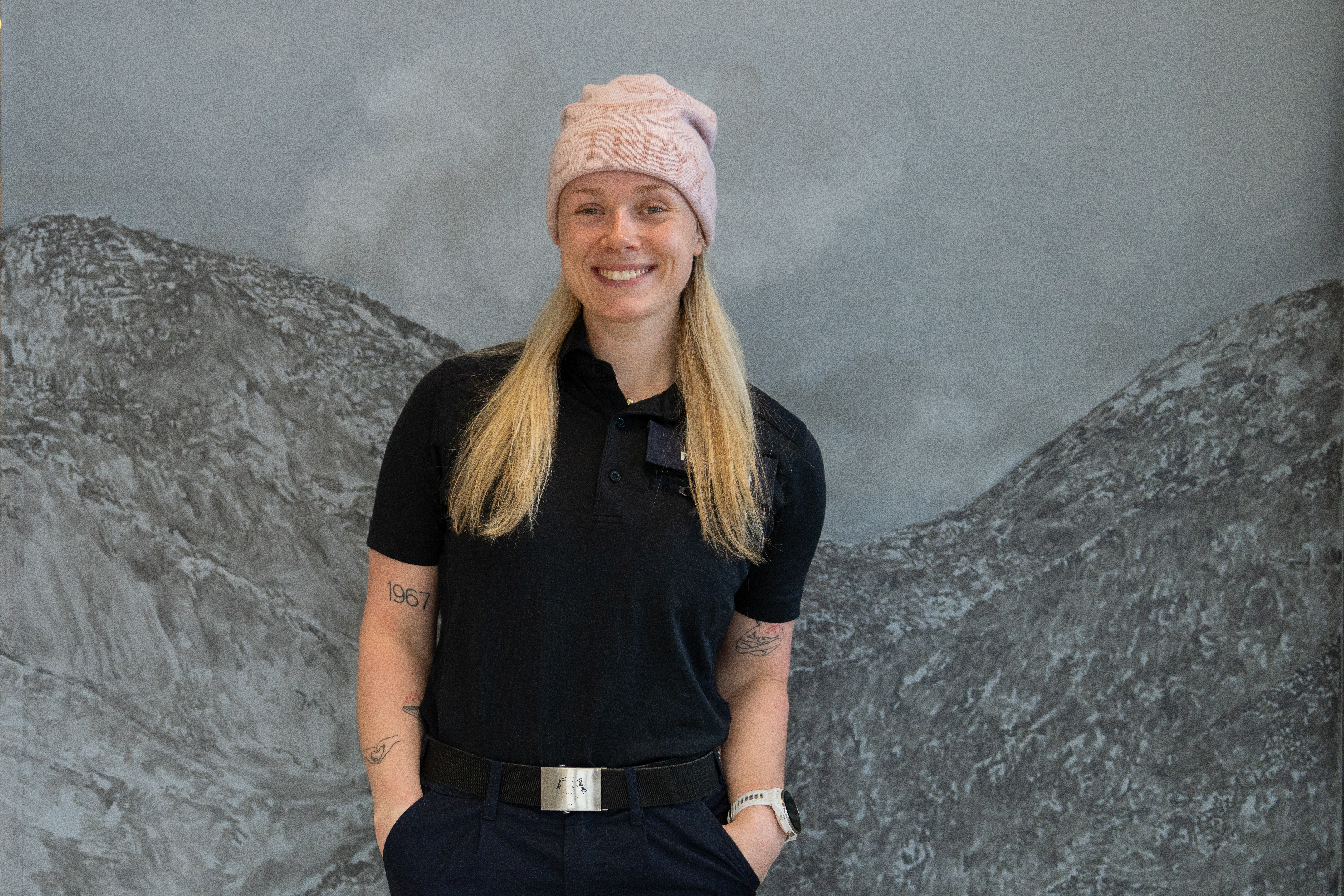 Kvinnelig student i uniform og rosa lue står foran en grå vegg. Veggen har illustrasjoner av fjell