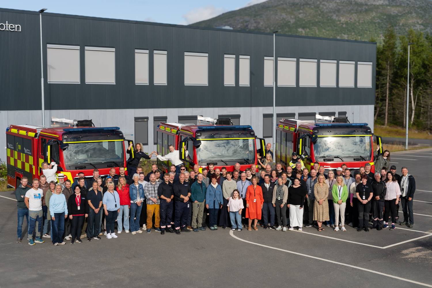 En stor gruppe mennesker står foran tre brannbiler og smiler mot kamera. Foto