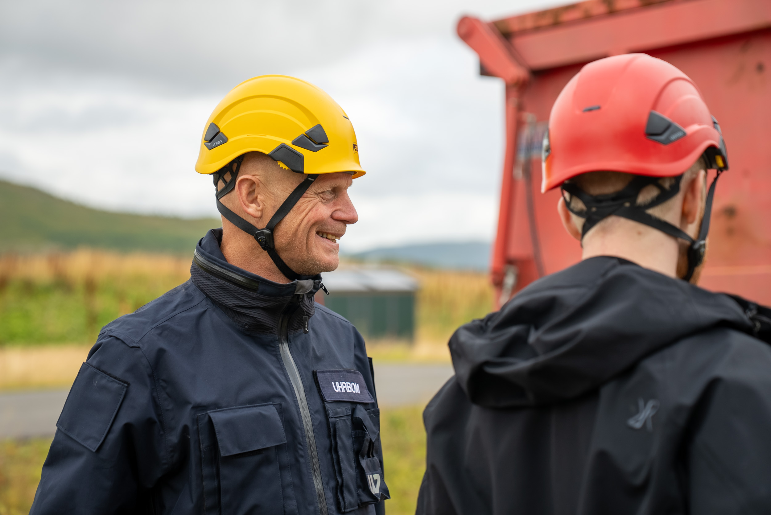 En mann med gul hjelm sett fra siden, en mann med rød hjelm med ryggen til. 