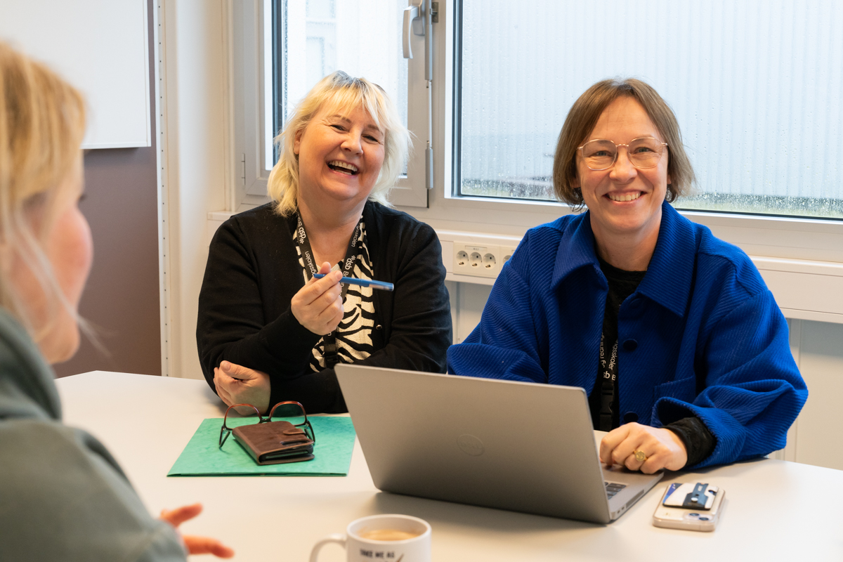 To smilende kvinner sitter ved et møtebord. Den ene har pc foran seg. Foto