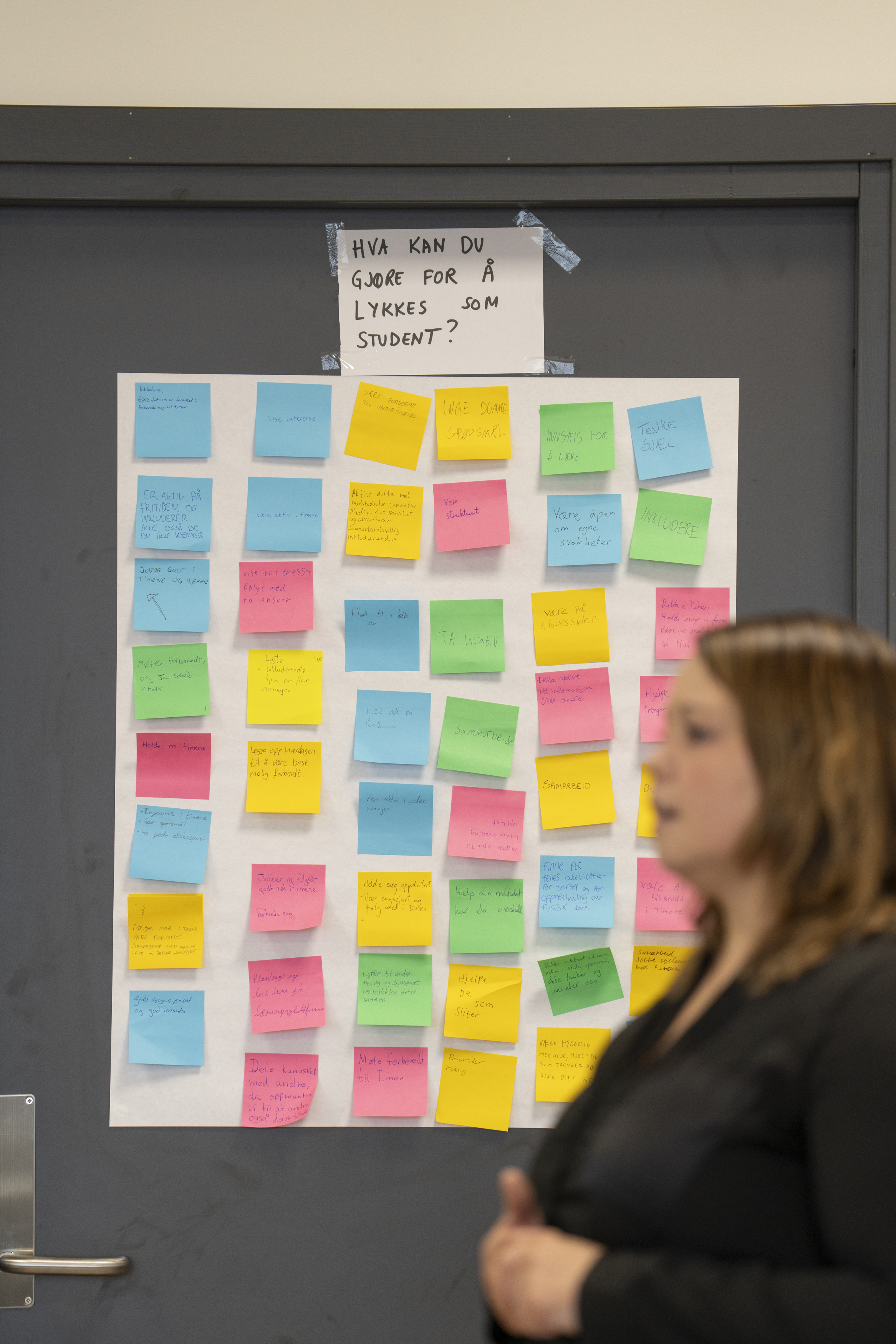 En kvinne står foran en dør som er dekket med post-it-lapper i ulike farger. Foto