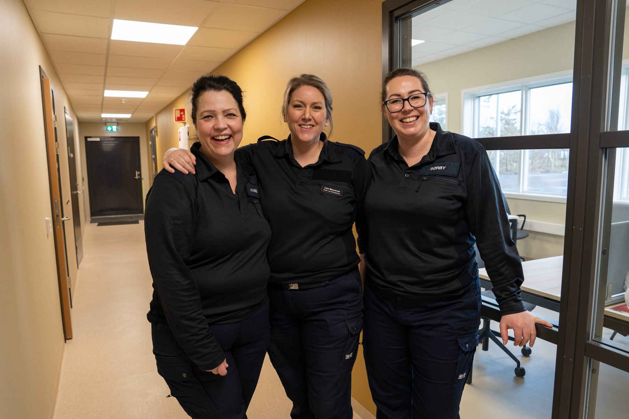 Tre smilende kvinner i sort uniform som står i en gang og holder rundt hverandre. Foto