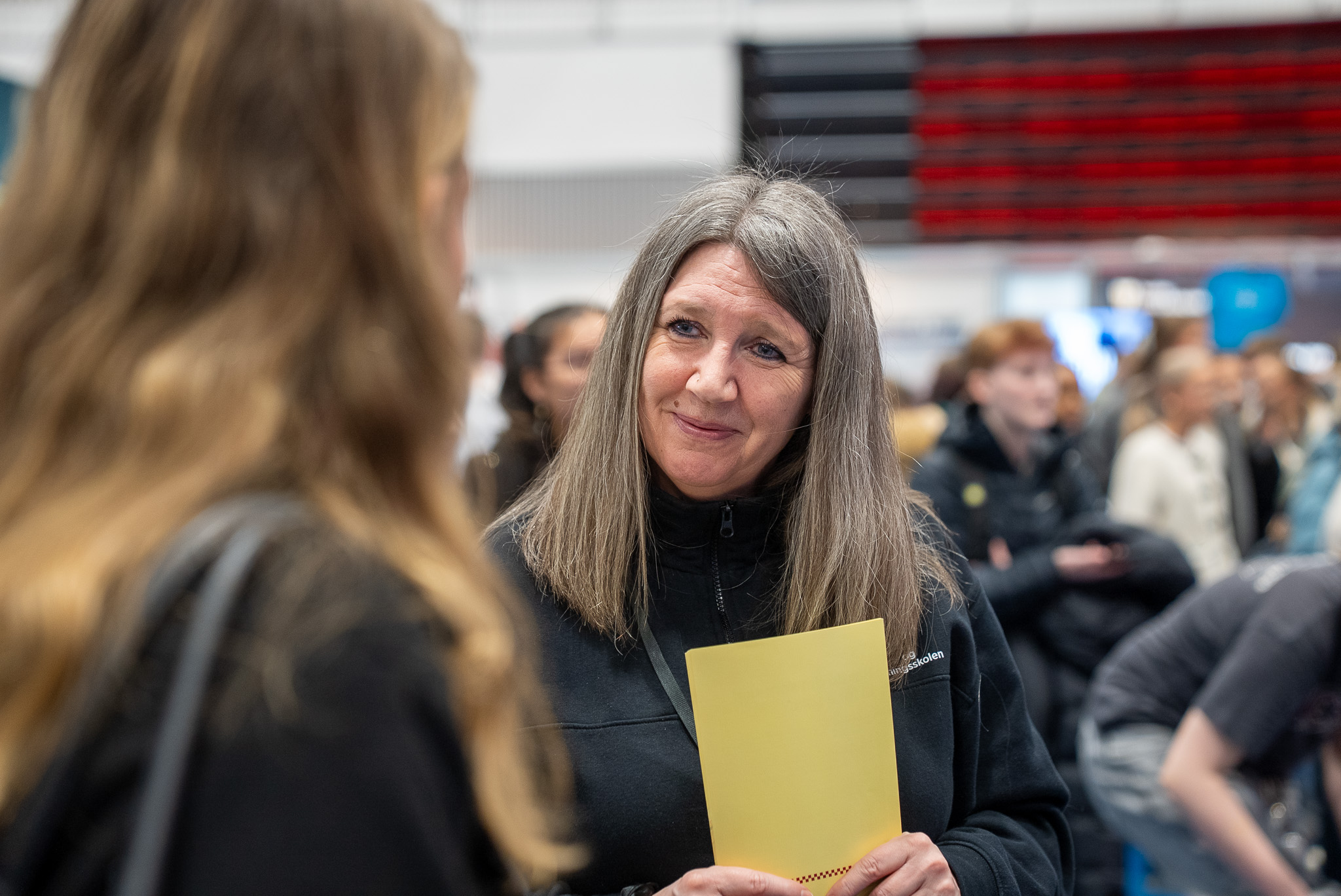 Nærbilde av en smilende kvinne med en brosjyre i hånda. Hun lytter til en elev. Foto. 