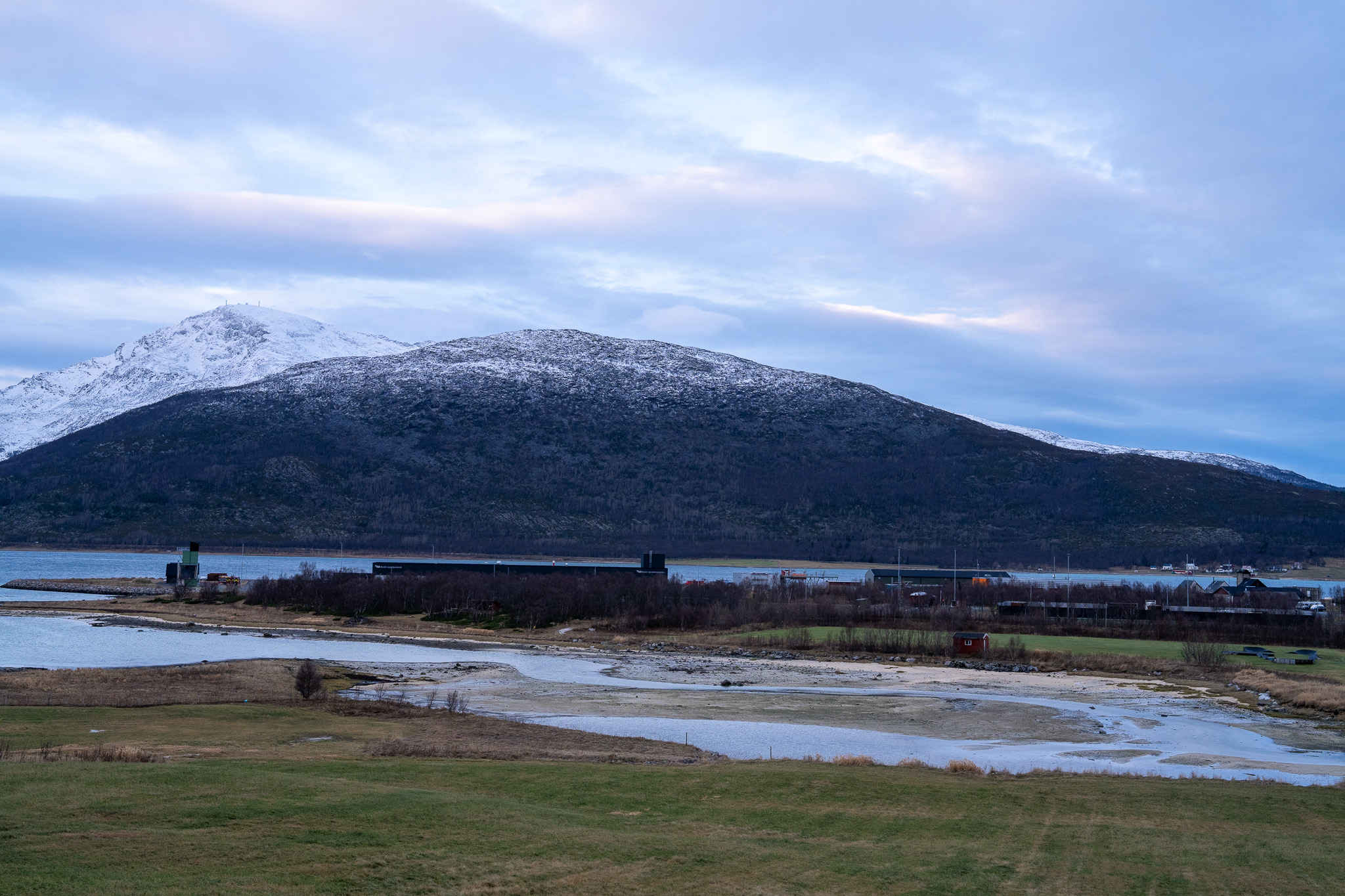 Oversiktsbilde med gress foran og fjord og fjell bak.