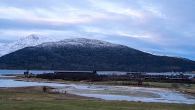 Oversiktsbilde med gress foran og fjord og fjell bak.