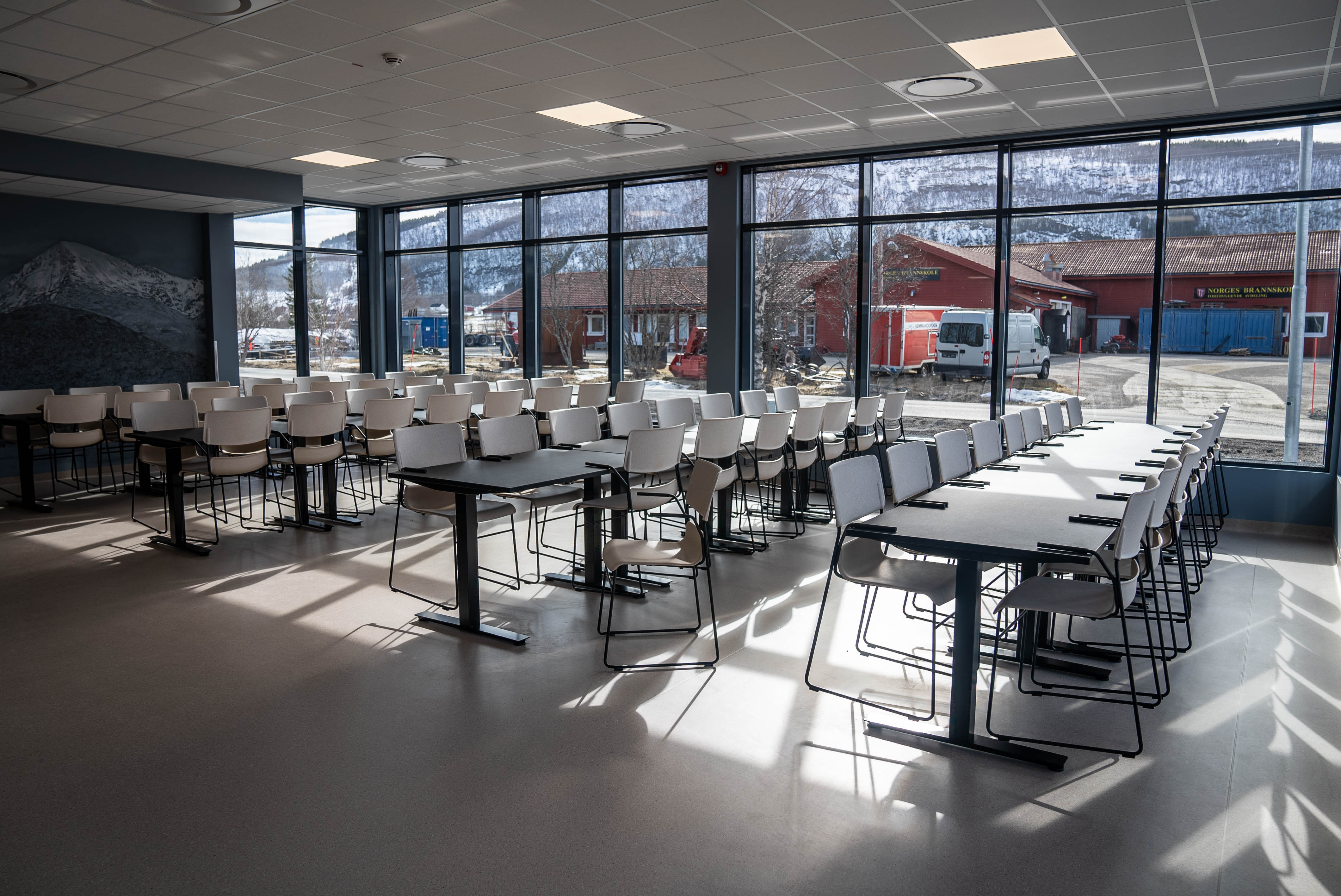 Tomme bord og stoler i en kantine. I bakgrunnen er store vinduer som slipper inn lys. Foto