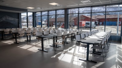 Tomme bord og stoler i en kantine. I bakgrunnen er store vinduer som slipper inn lys. Foto