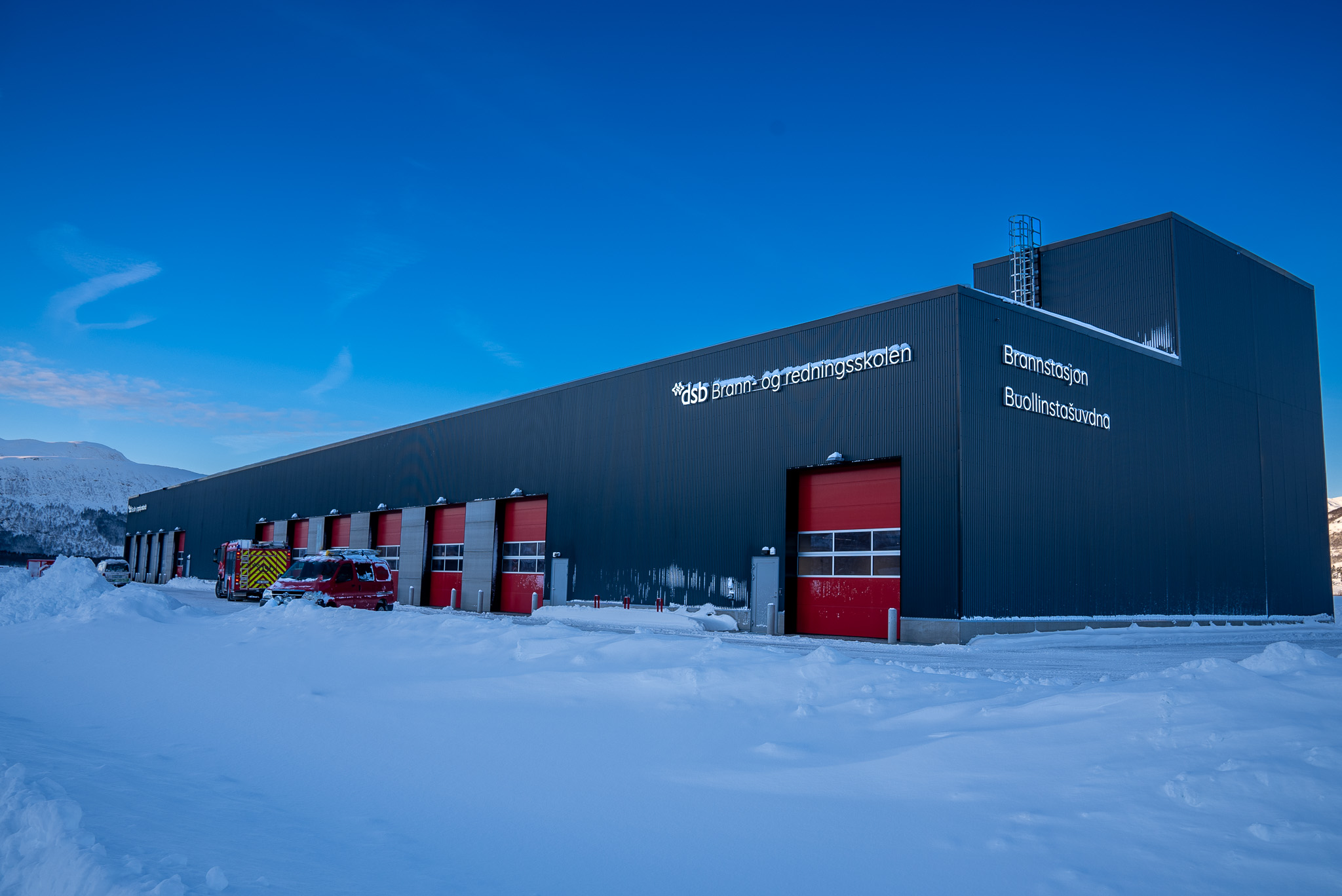 En brannstasjon med røde dører og snø på bakken foran. Himmelen bak er knallblå