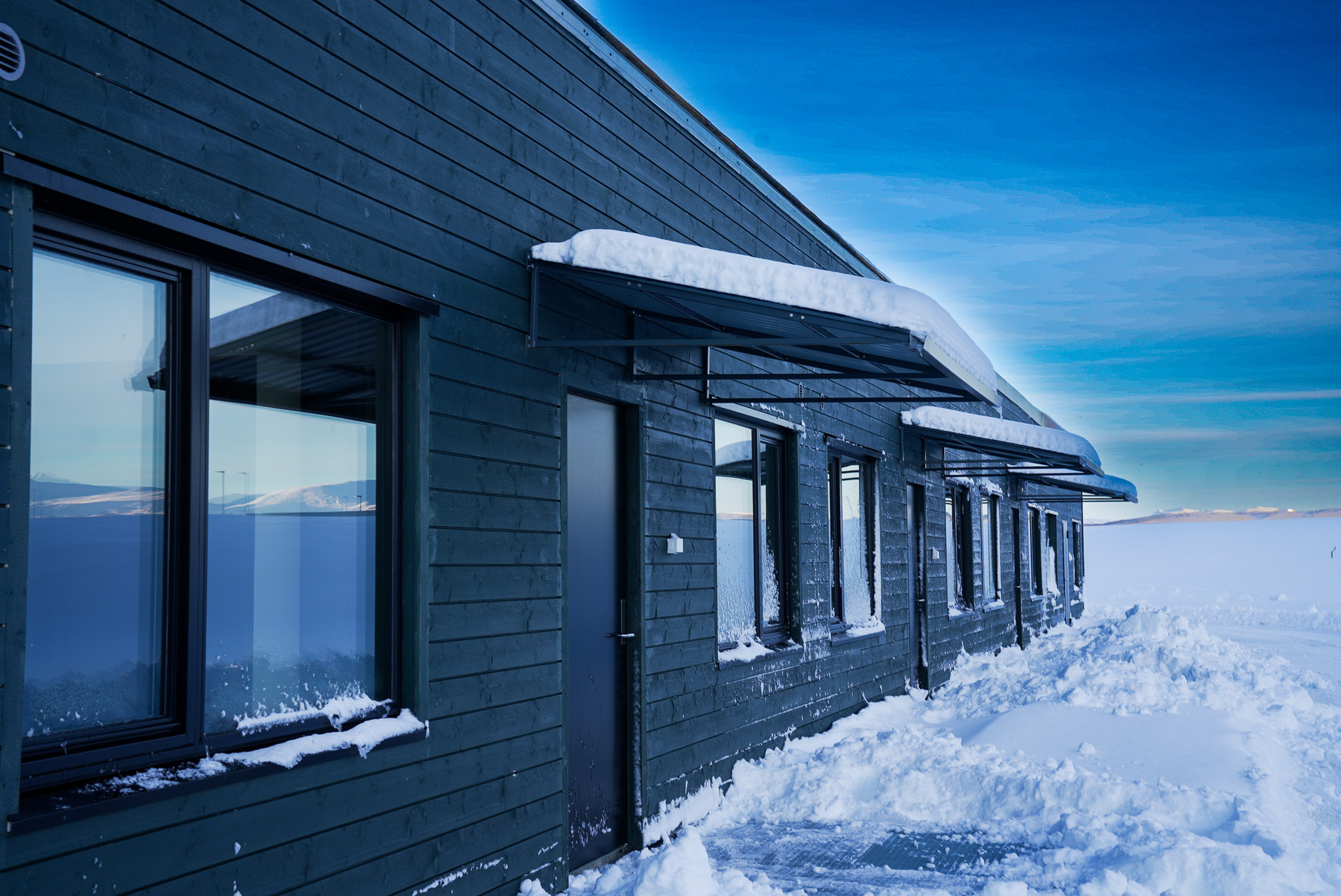Fire hus i rekke, blå himmel bak. Snø på bakken foran. 