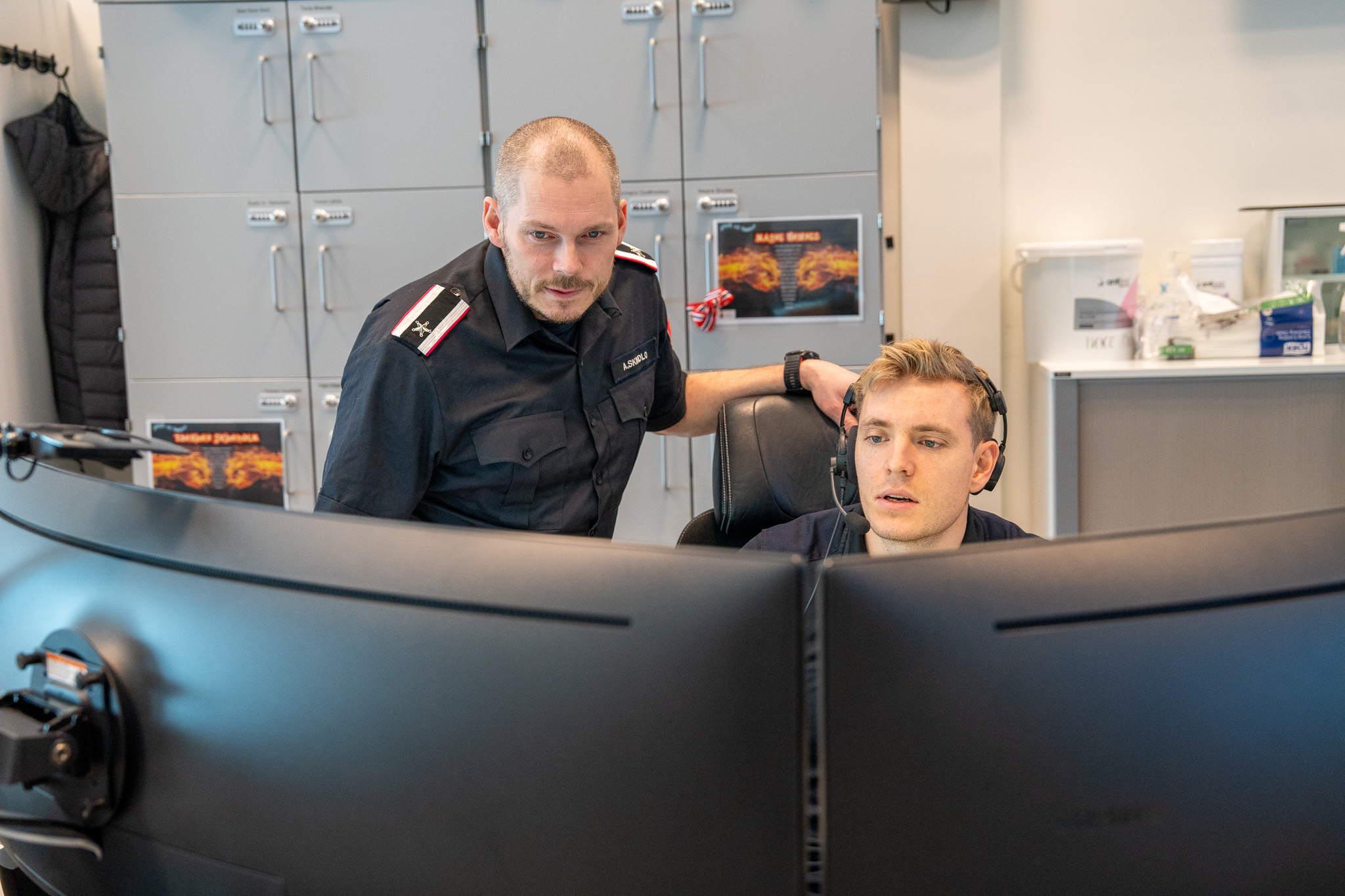 To mannlige operatører sitter bak skjermer på en 110-sentral