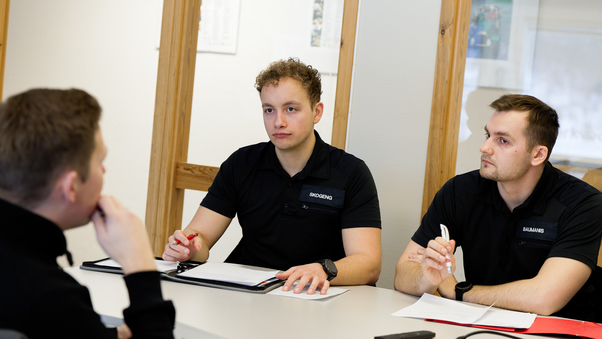 To menn i uniform sitter med blyant og notatblokk, i samtale med en mann med ryggen til kamera