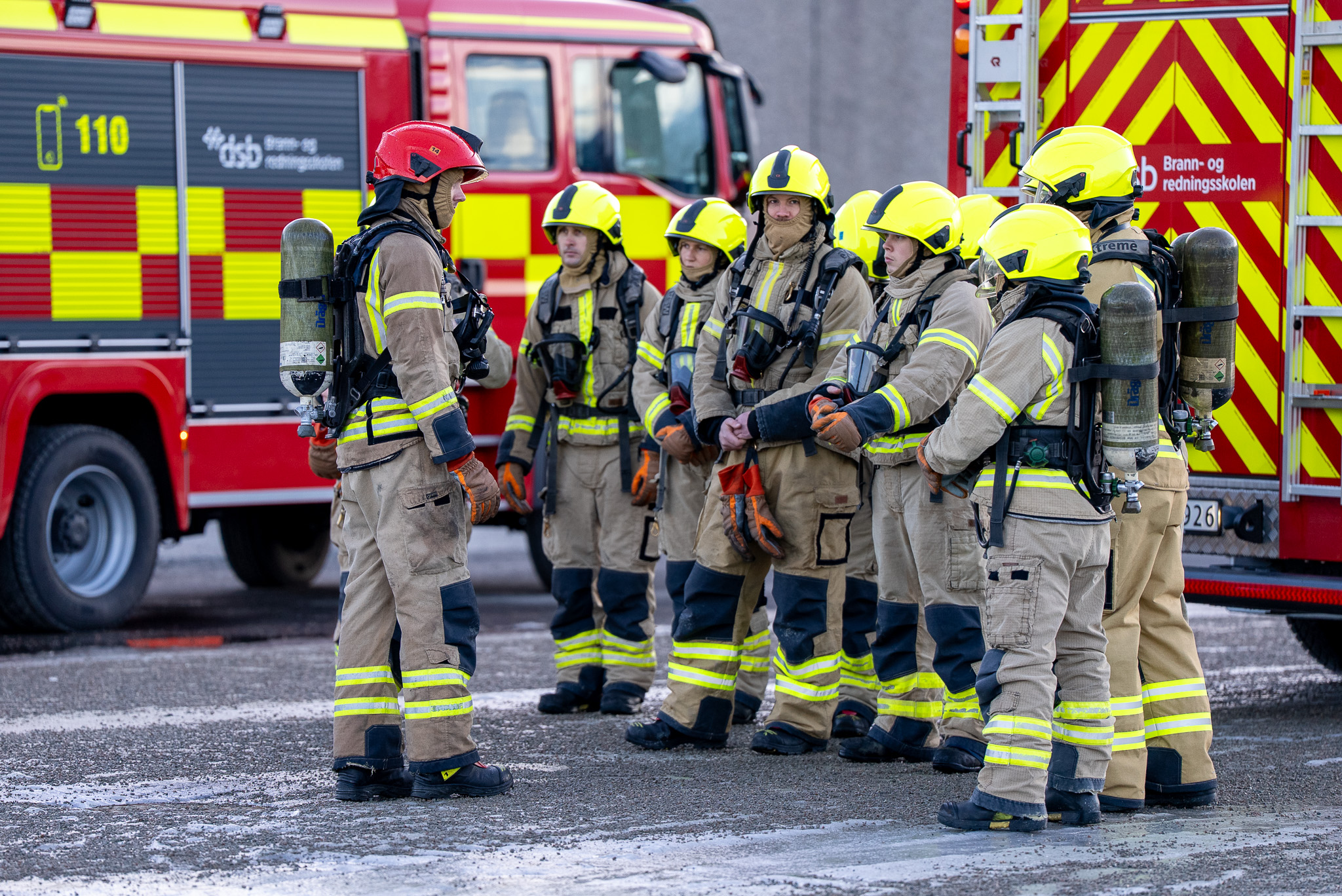 Studenter i røykdykkeruniform som får undervisning ute på felt. I bakgrunn er det to brannbilder.