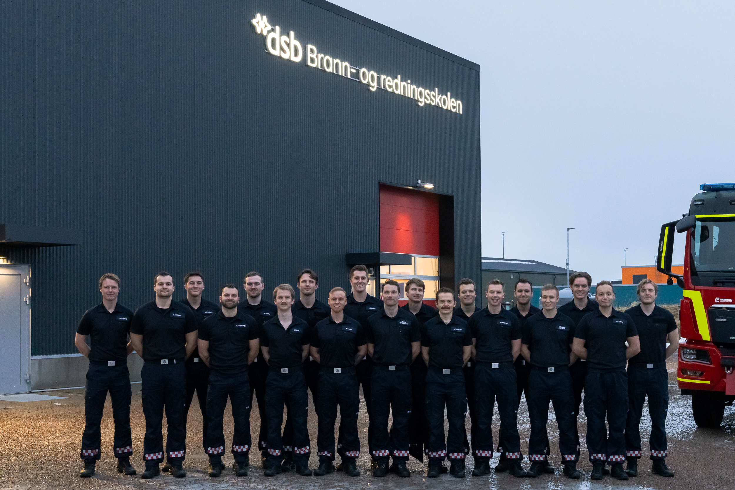 En gruppe studenter står oppstilt i uniform foran en brannstasjon