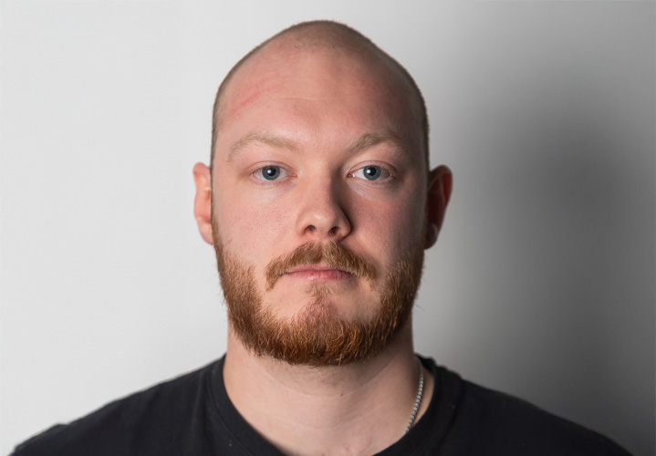 Ung mann med skjegg og bart ser inn i kamera. Portrettbilde