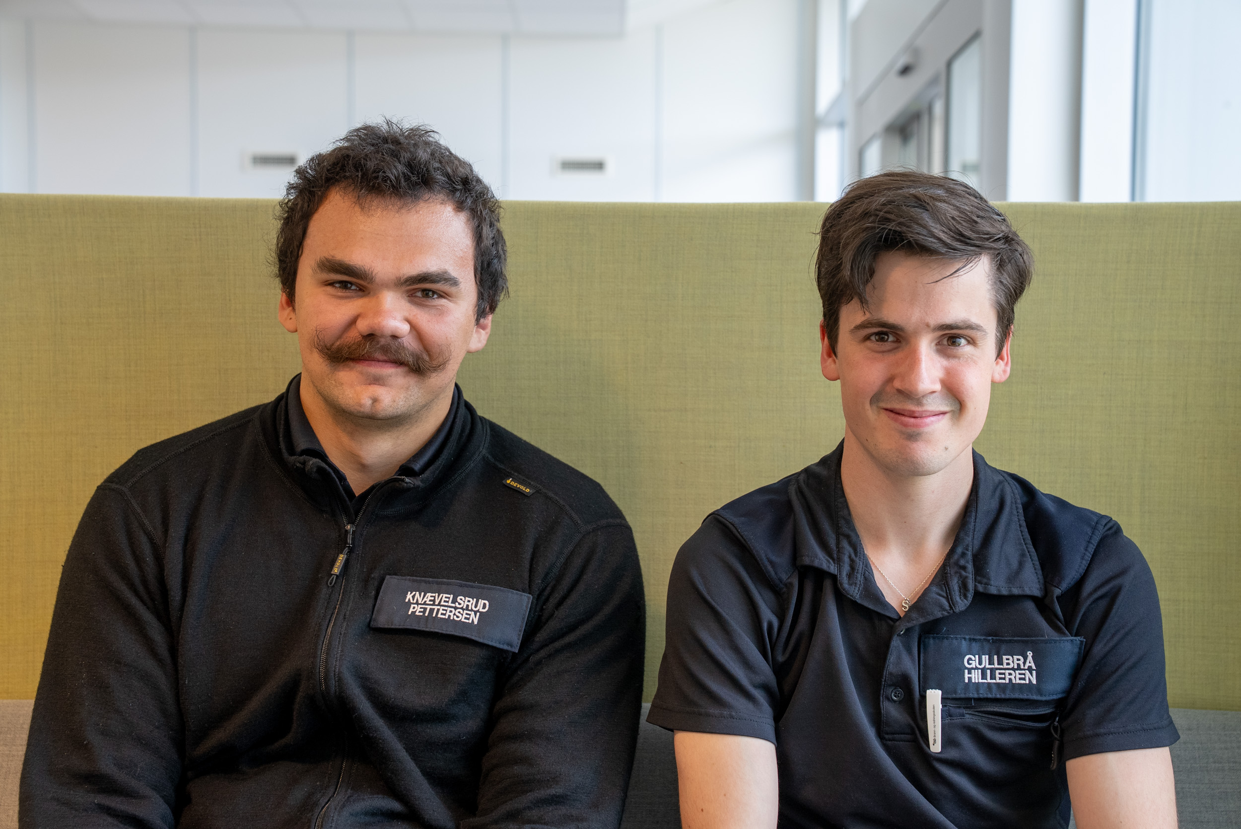 To menn i sort uniform sitter i en grønn sofa og smiler mot kamera. Foto