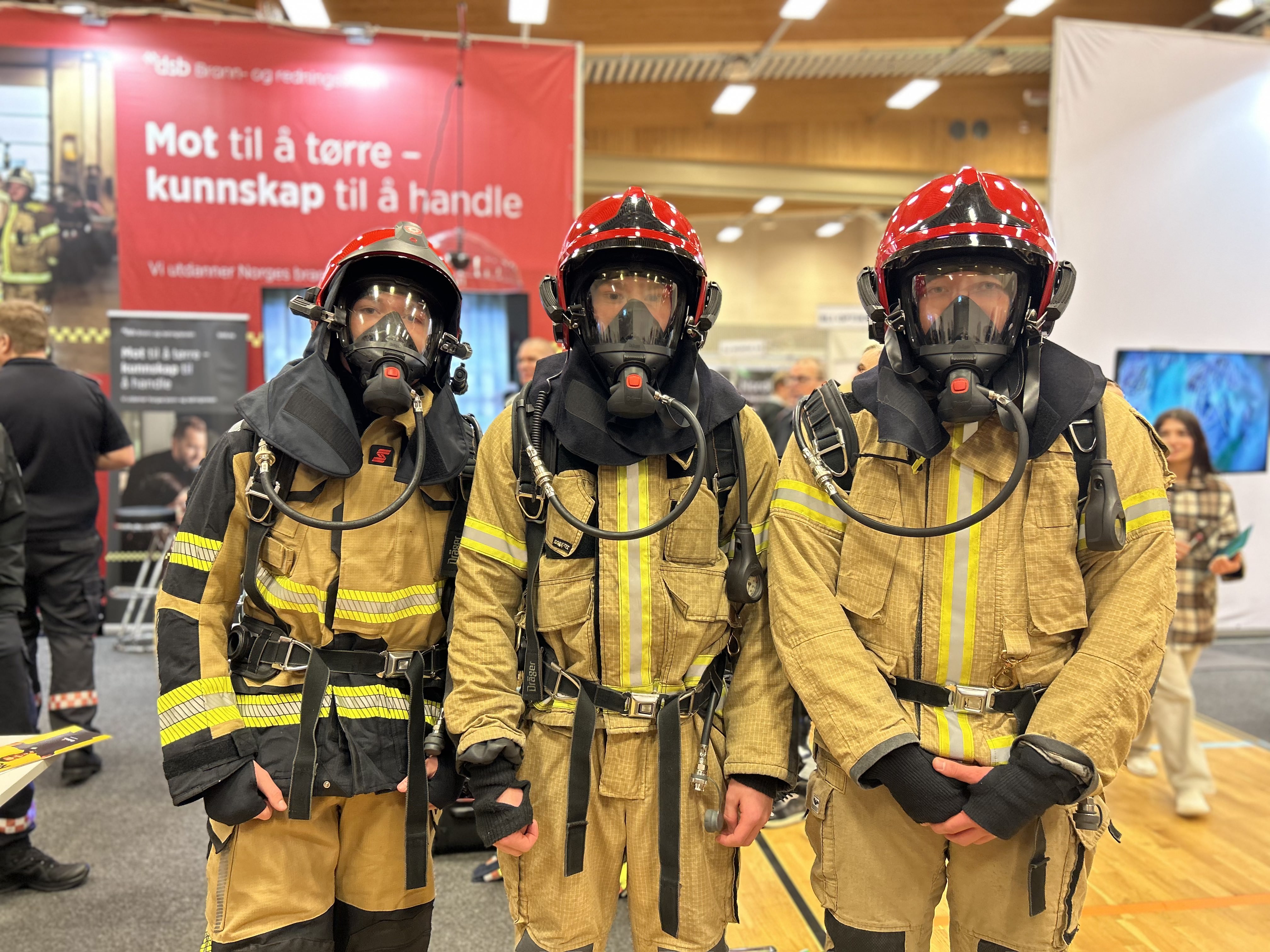 Tre elever i full røykdykkerbekledning, inkludert masker, står oppstilt mot kamera