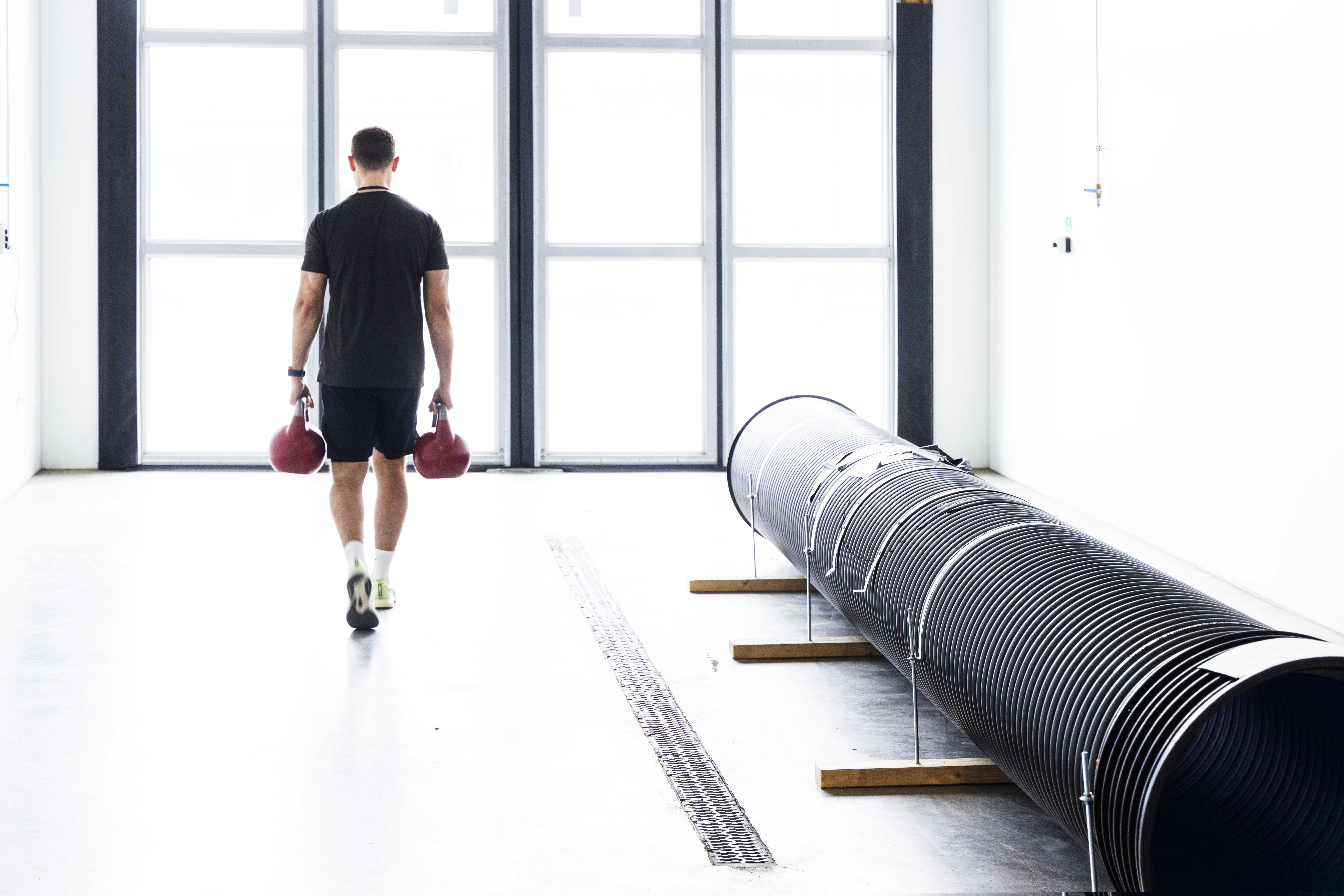 En mann går med en kettlebell i hver hånd med ryggen mot kamera