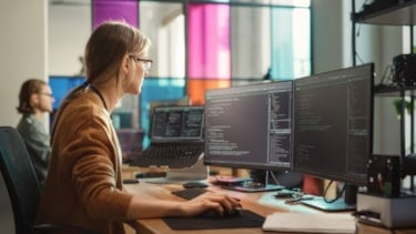 Kvinne sitter og programmerer på pc. I bakgrunnen sitter en mann og gjør det samme