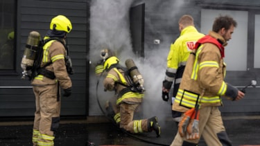 Tre brannkonstabler og en intstruktør. Den ene brannkonstabelen er på vei inn ei dør med røyk som kommer ut.