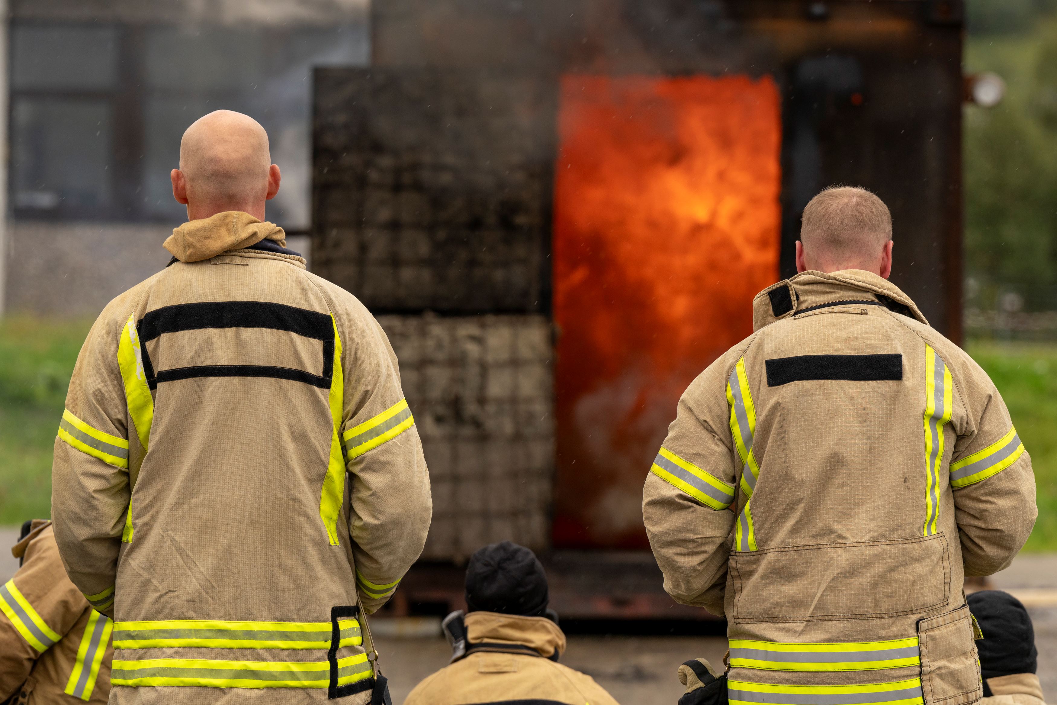 To brannkonstabler står med ryggen mot kamera og observerer container som brenner. Foto