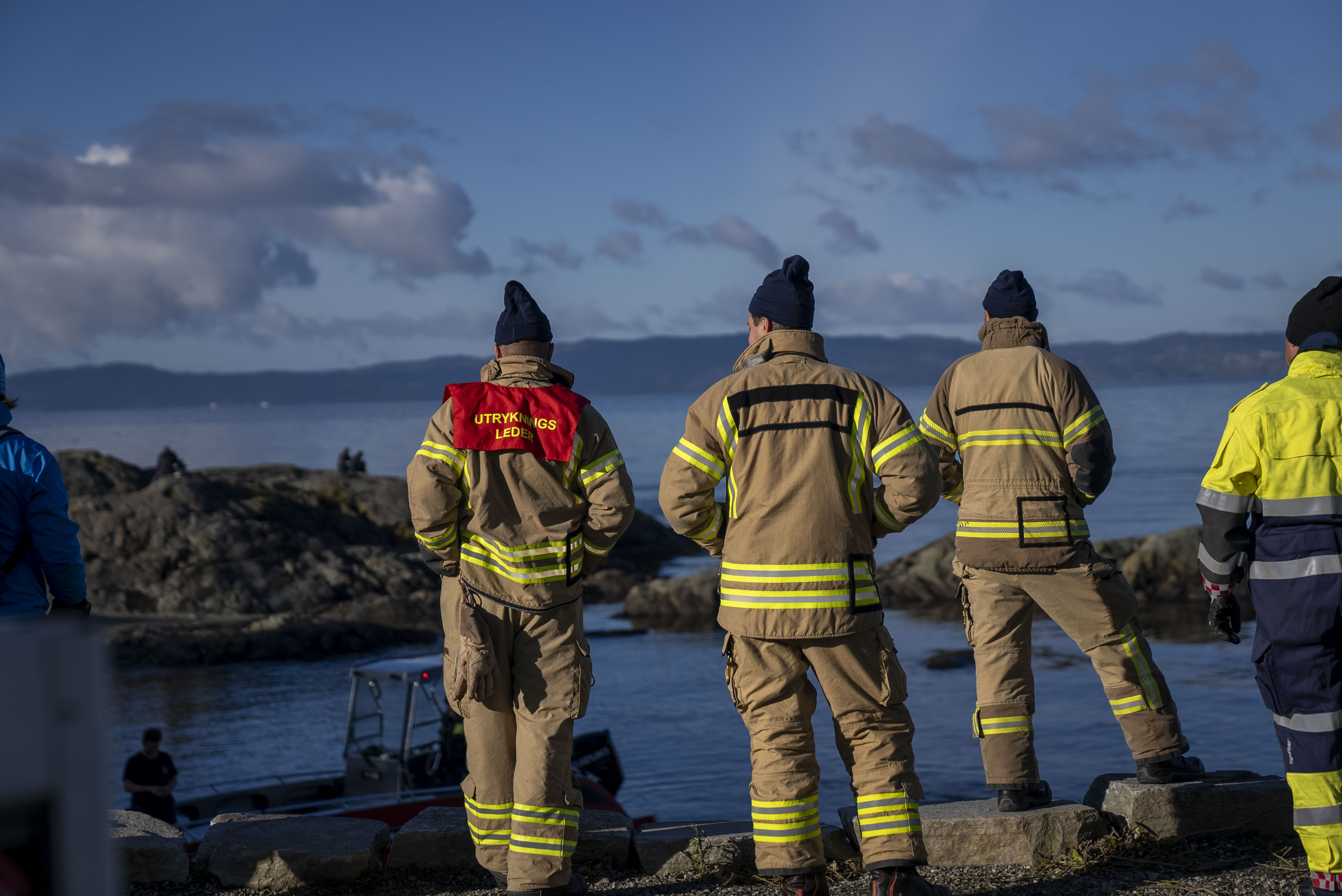 Tre menn i brannbekledning står med ryggen til og speider ut mot fjorden
